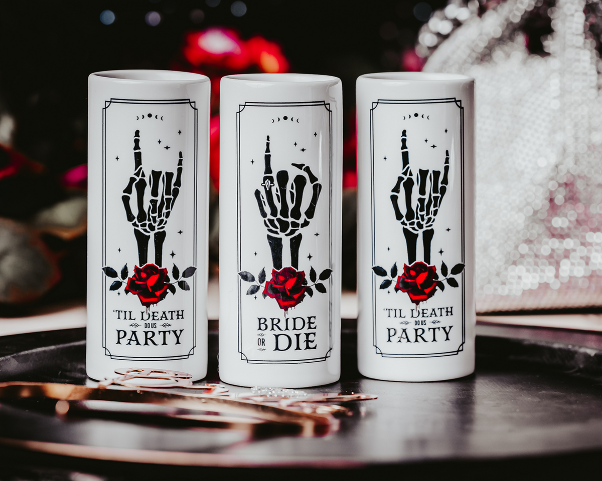 Gothic-style ceramic shot glasses featuring skeleton hand designs with red roses and phrases like &quot;Bride or Die&quot; and &quot;&#39;Til Death Do Us Party,&quot; perfect for an alternative bridal party.