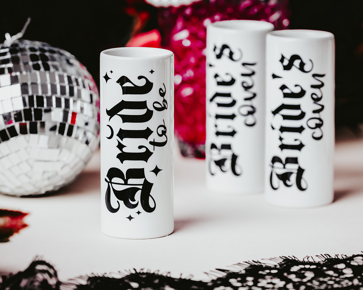 Ceramic shot glasses with gothic-style &#39;Bride to Be&#39; and &#39;Bride&#39;s Coven&#39; typography, perfect for a non-traditional bride and her bridal party, set against a dark, elegant backdrop.