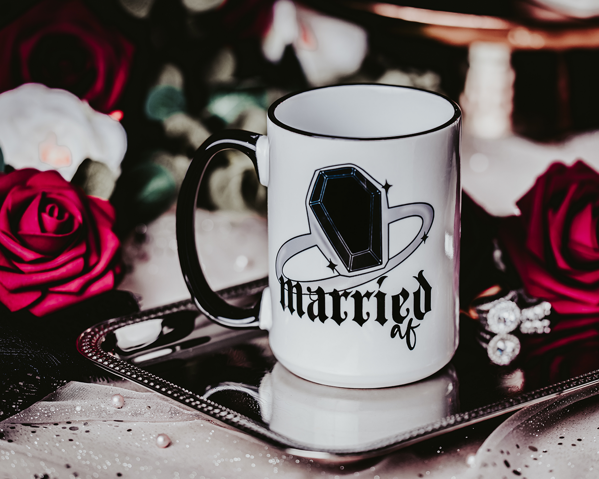 White ceramic mug with a black rim and handle, featuring the phrase 'Married AF' in bold, gothic lettering along with a coffin ring illustration. The mug is set on a black tray with a crystal accent, surrounded by a soft, romantic background of red roses and other decorative elements, capturing a gothic bridal theme.