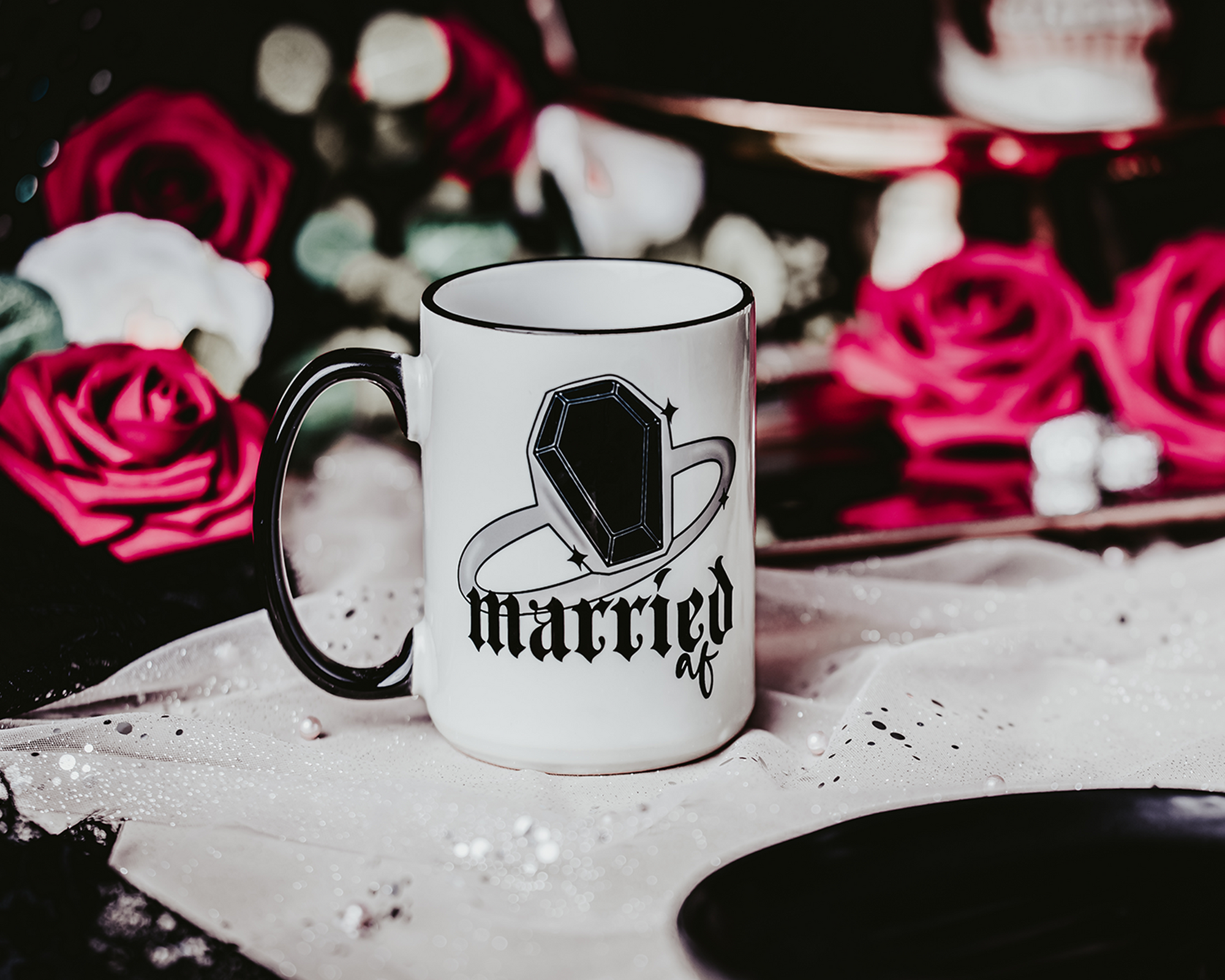 White ceramic mug with a black rim and handle, featuring the phrase 'Married AF' in bold, gothic lettering along with a coffin ring illustration. The mug is set on a black tray with a crystal accent, surrounded by a soft, romantic background of red roses and other decorative elements, capturing a gothic bridal theme.