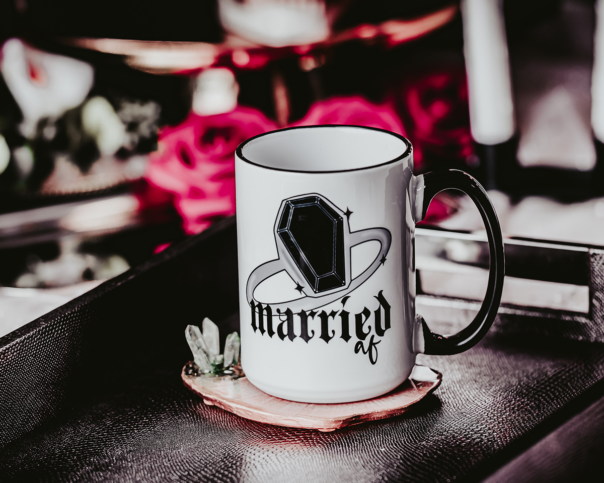 White ceramic mug with a black rim and handle, featuring the phrase &#39;Married AF&#39; in bold, gothic lettering along with a coffin ring illustration. The mug is set on a black tray with a crystal accent, surrounded by a soft, romantic background of red roses and other decorative elements, capturing a gothic bridal theme.