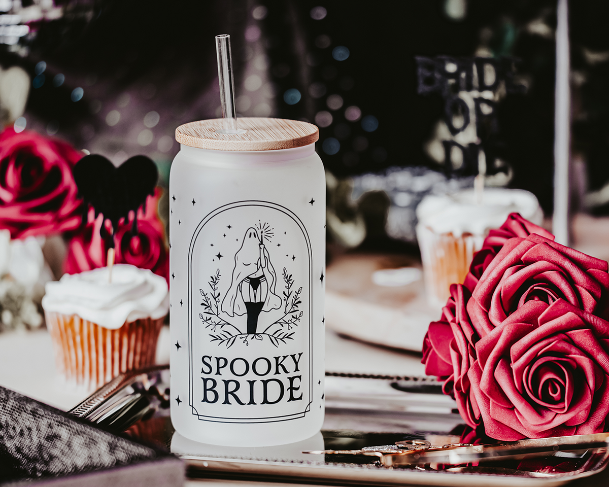 Spooky Bride Frosted Glass Can Tumbler featuring a female ghost design in stockings, with a bamboo lid and clear reusable straw, set against a gothic-themed backdrop with roses and dark décor elements.&quot;